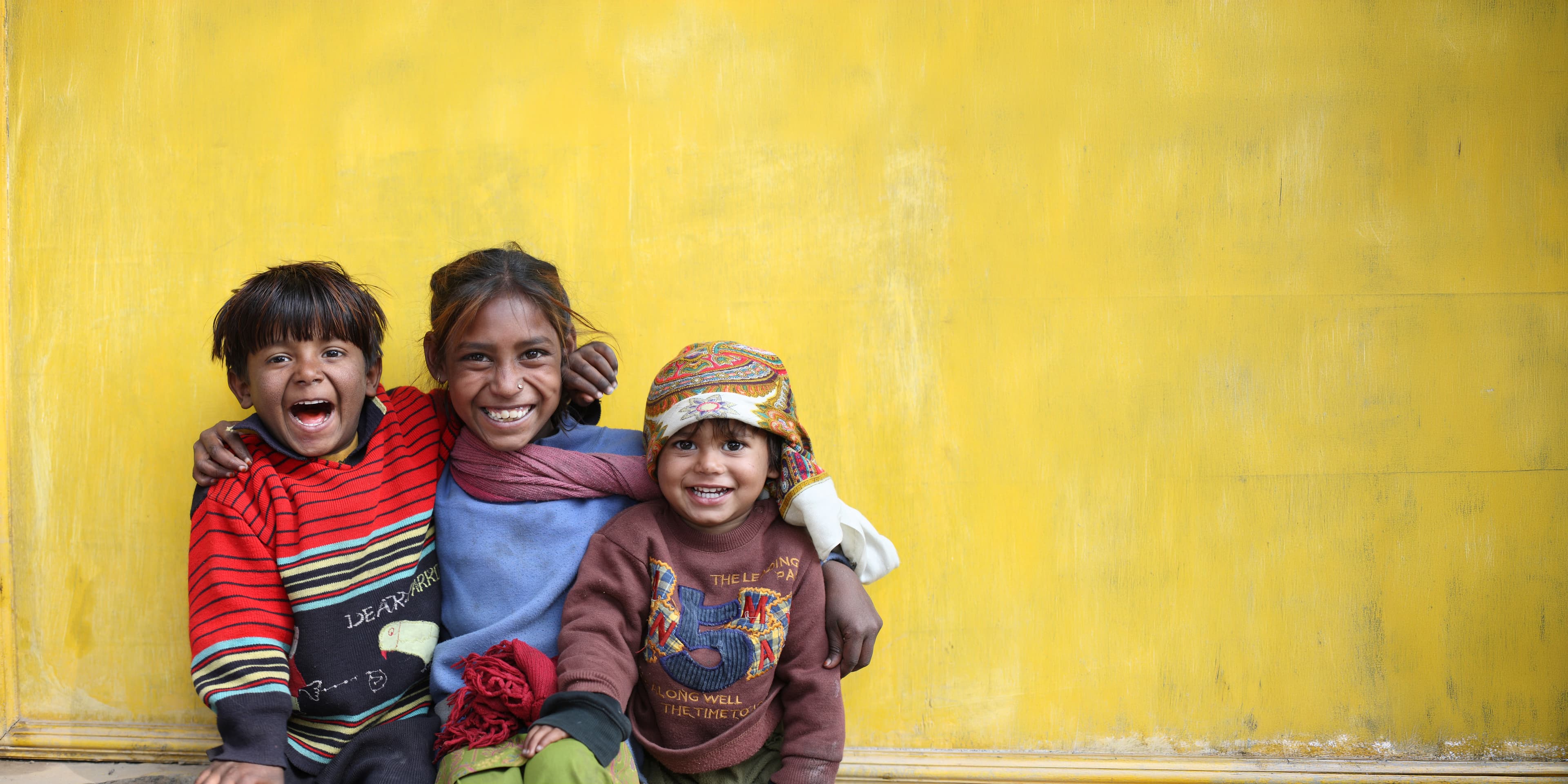Three children smiling against a yellow wall in Nepal